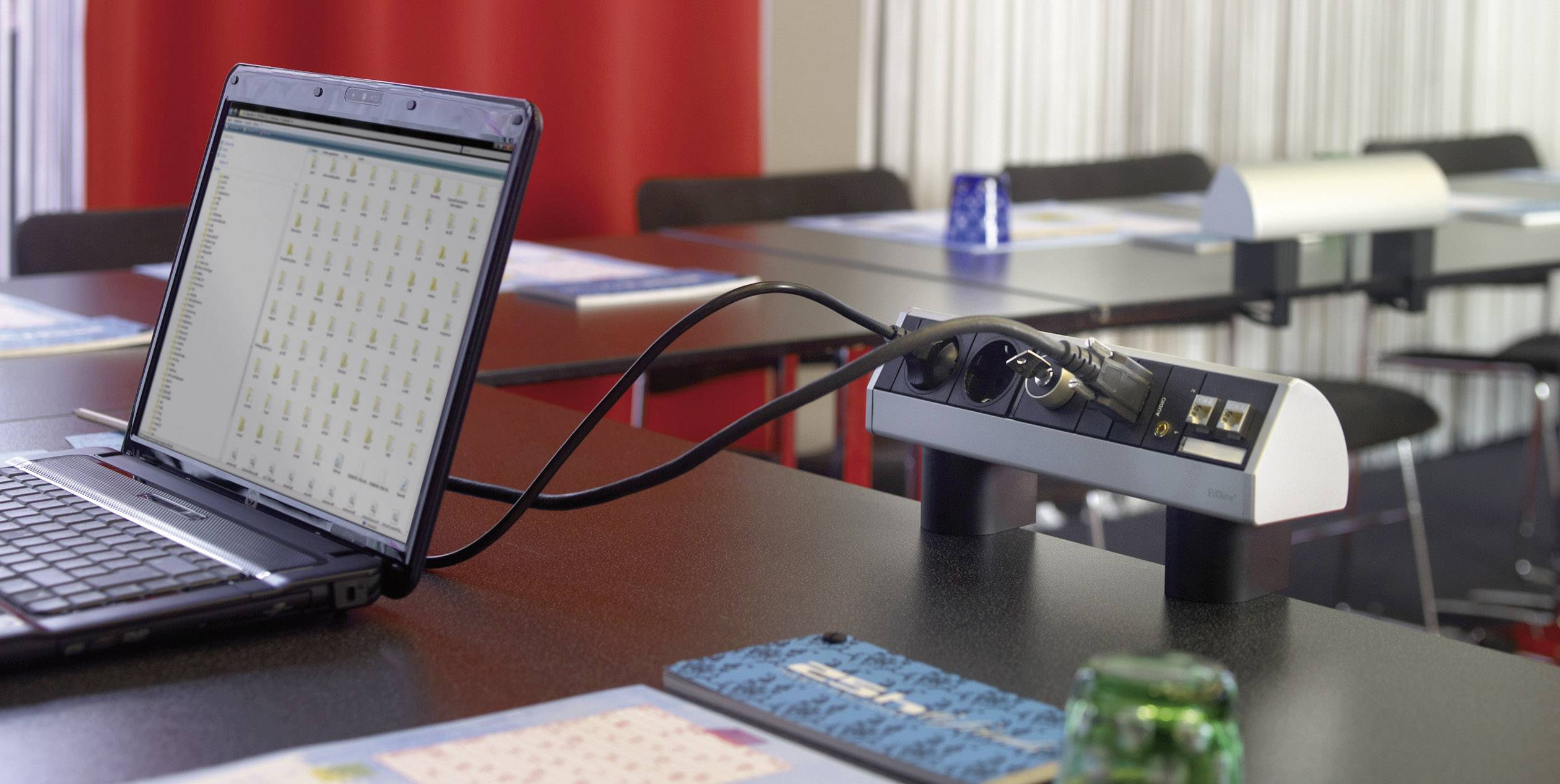 A laptop is connected on a table in a meeting room. The table is equipped with multiple power strips.
