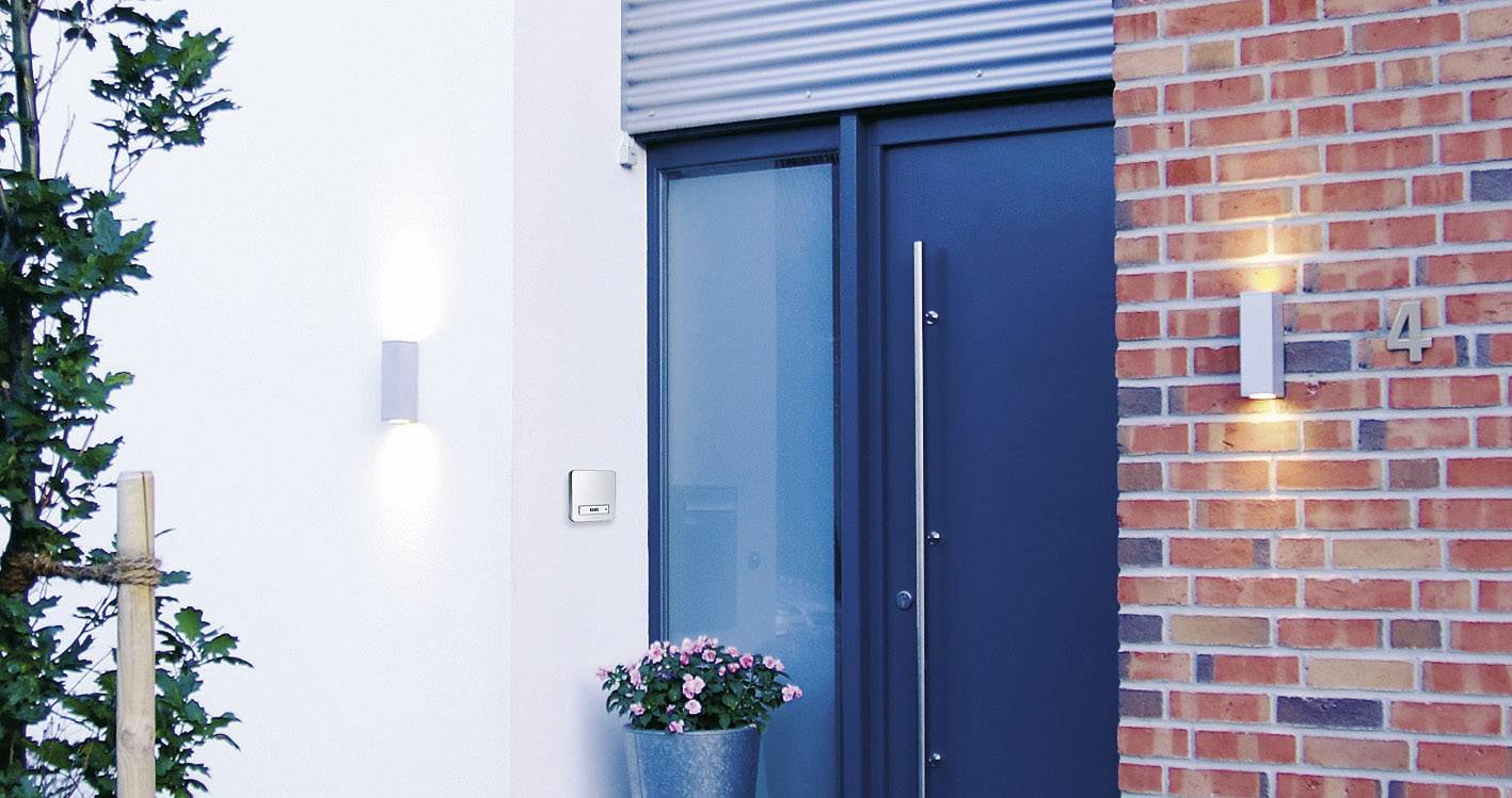 A modern entrance door in blue, surrounded by brick and white walls. A pot with pink flowers stands to the left of the door.
