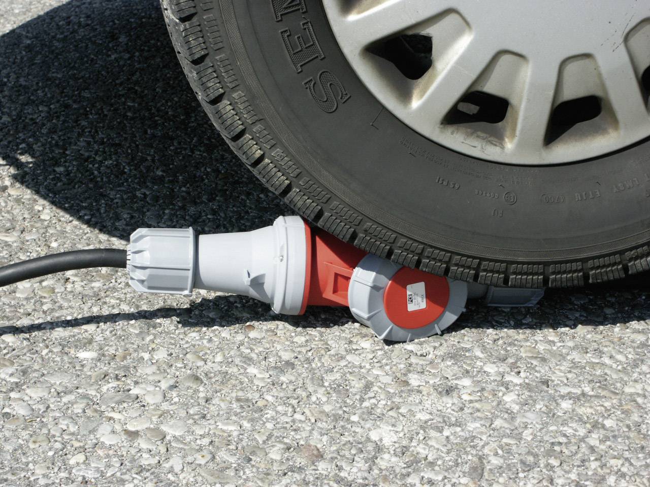 A car tyre sits on a connector on an asphalt road. The connector is red and grey and is pinched by the tyre.