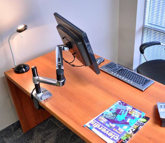 A modern desk with a monitor mount, screen, keyboard, lamp and chair. A technology magazine is lying on the table.