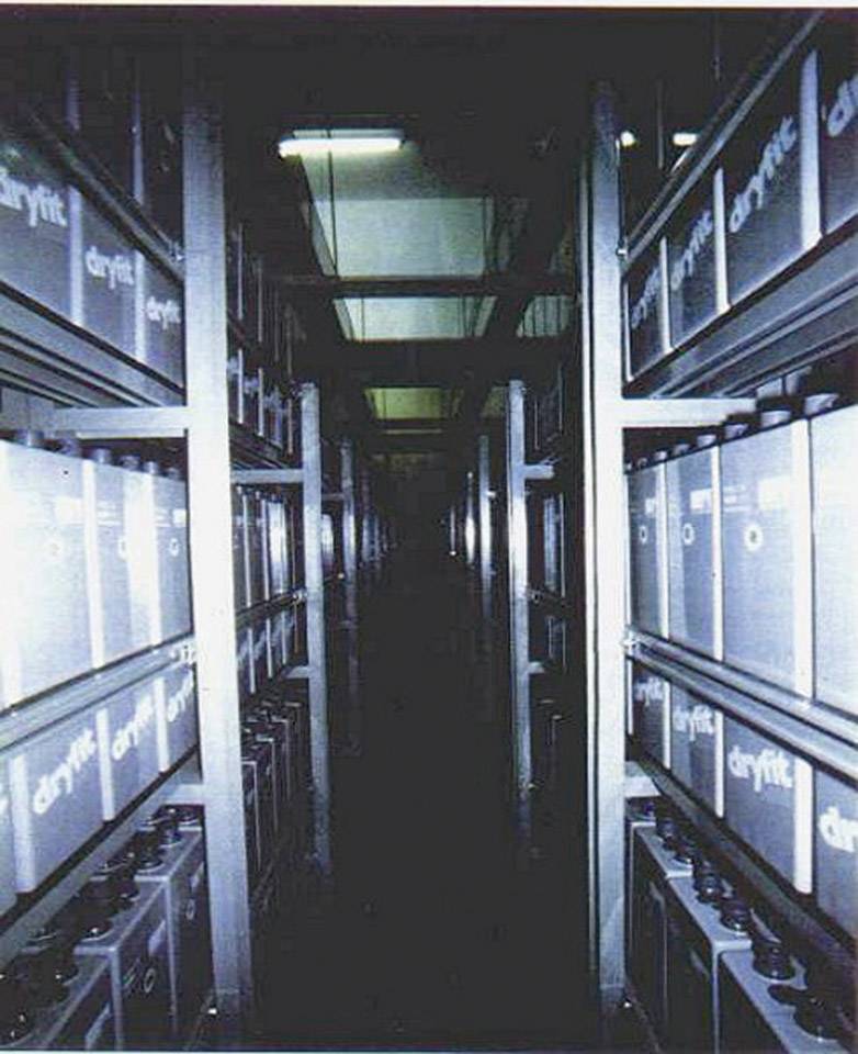 A long, narrow corridor between towering shelving units with large canisters bearing the word 'dryptik' in a dimly lit room.
