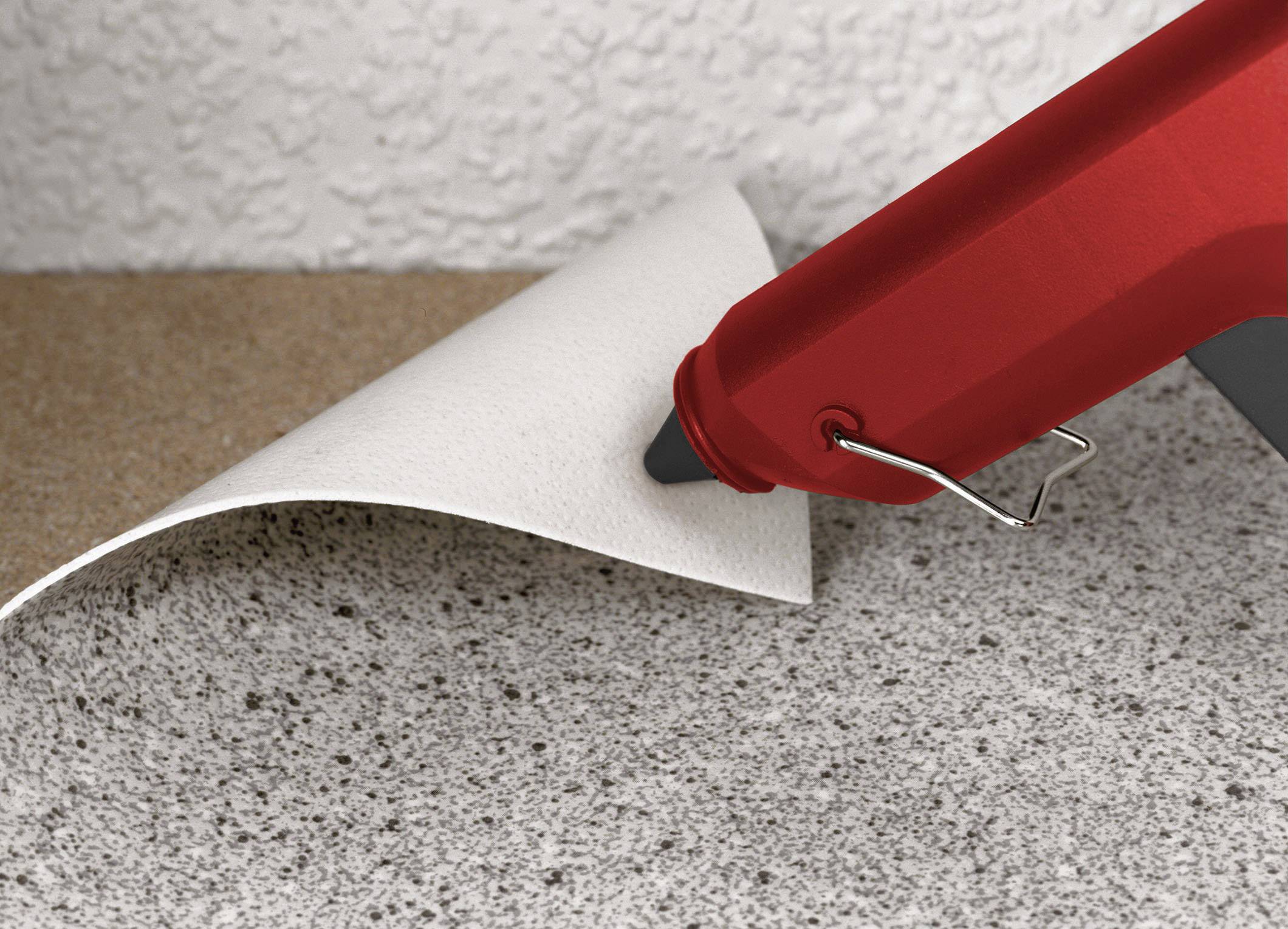 A red hot glue gun lifts the corner of a floor tile, revealing the adhesive underneath.