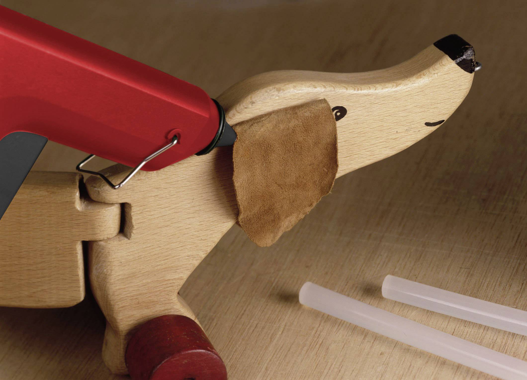 A red hot glue gun is sticking a brown ear to a wooden dog toy. Two glue sticks are lying beside it.