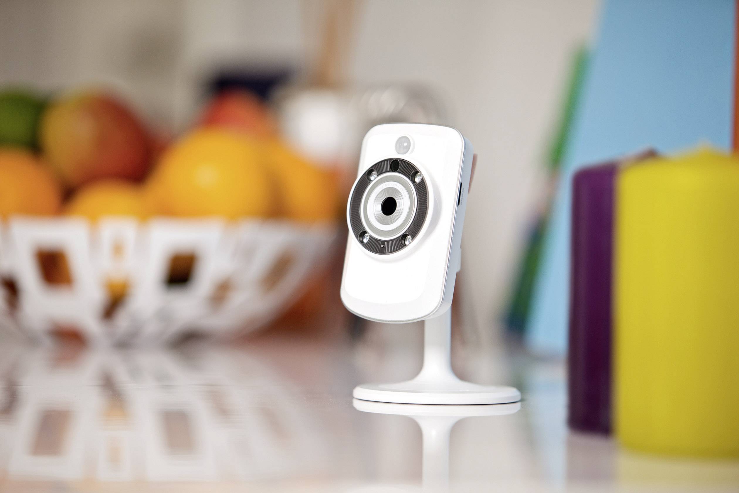 A small surveillance camera on a table in front of a blurred background with a fruit bowl and colourful folders.