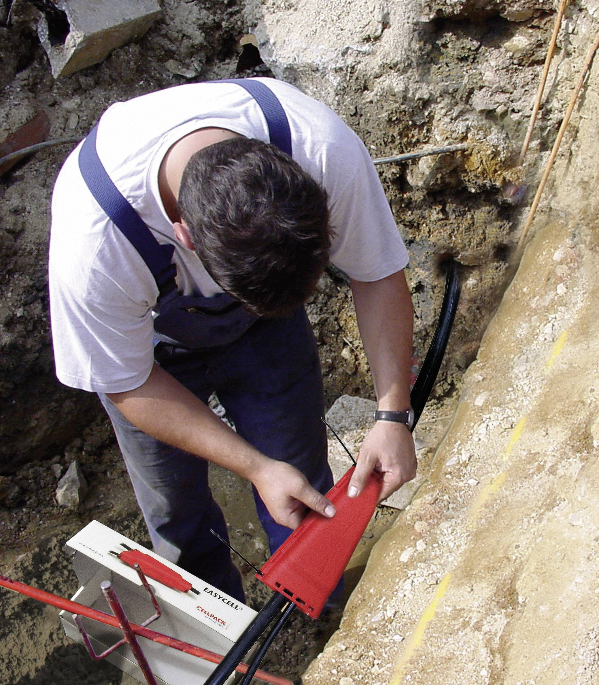 A worker in workwear is laying cables underground and inspecting a red protective conduit. Environmental conditions suggest construction work is in progress.