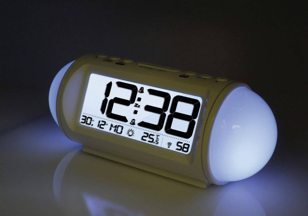 Digital alarm clock with illuminated display shows time as 12:38, date as 30/12 and temperature as 25.5 degrees. Background is dark.