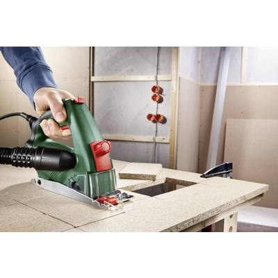 A person is operating a green electric saw and cutting a wooden board on a workbench in a workshop environment.
