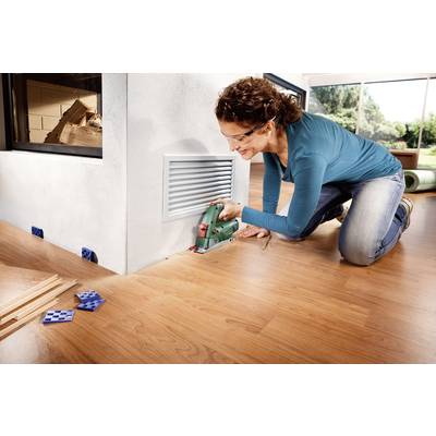 A woman is laying a laminate floor and cutting a piece to size with an electric jigsaw. Wooden strips and spacers are lying beside her.