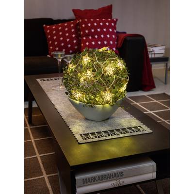 A plant pot wrapped with fairy lights sits on a table in a living room. Red cushions are visible in the background.