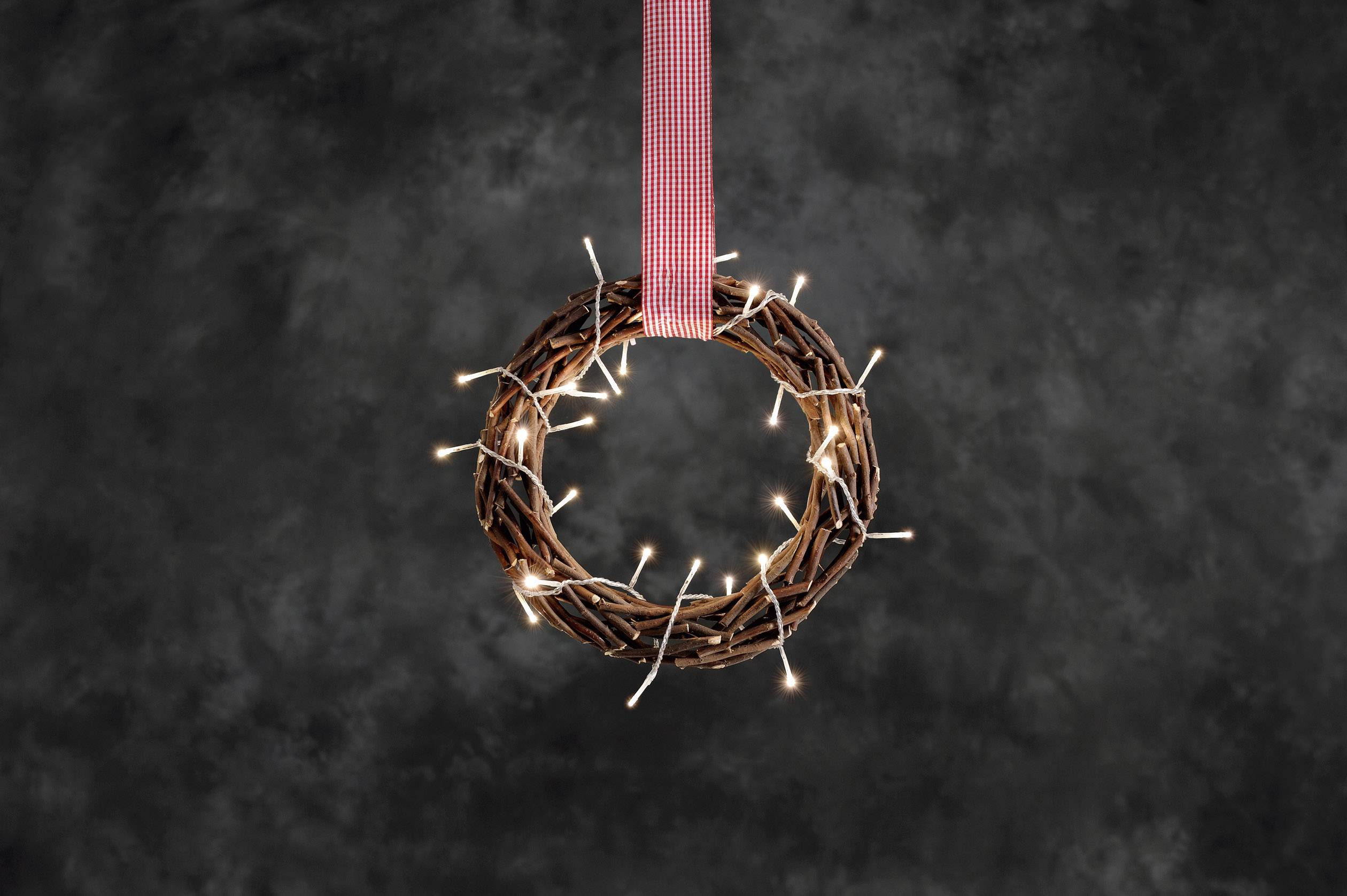 A wreath of branches with glowing lights, suspended from a red ribbon against a dark background.