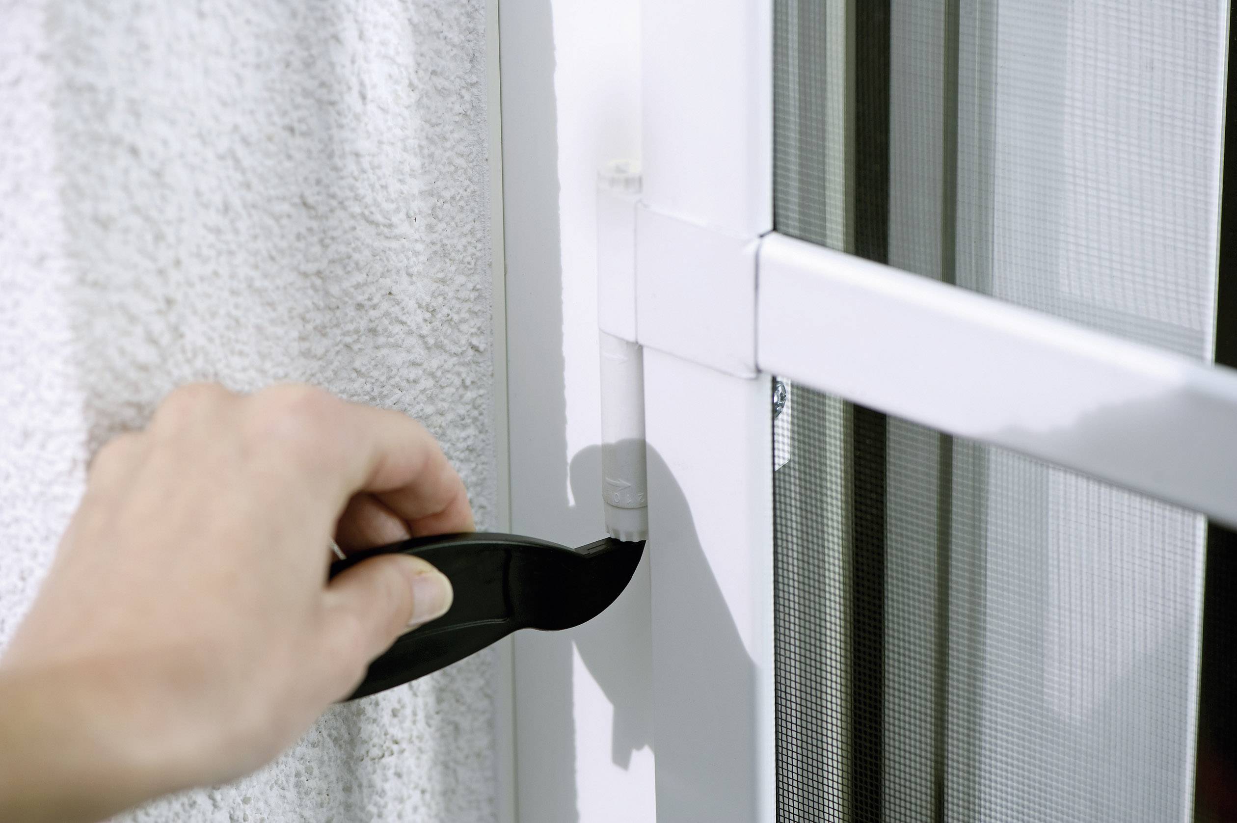 A person is cleaning the hinges of a white door with a black tool. The door has a glass window and a fly screen.