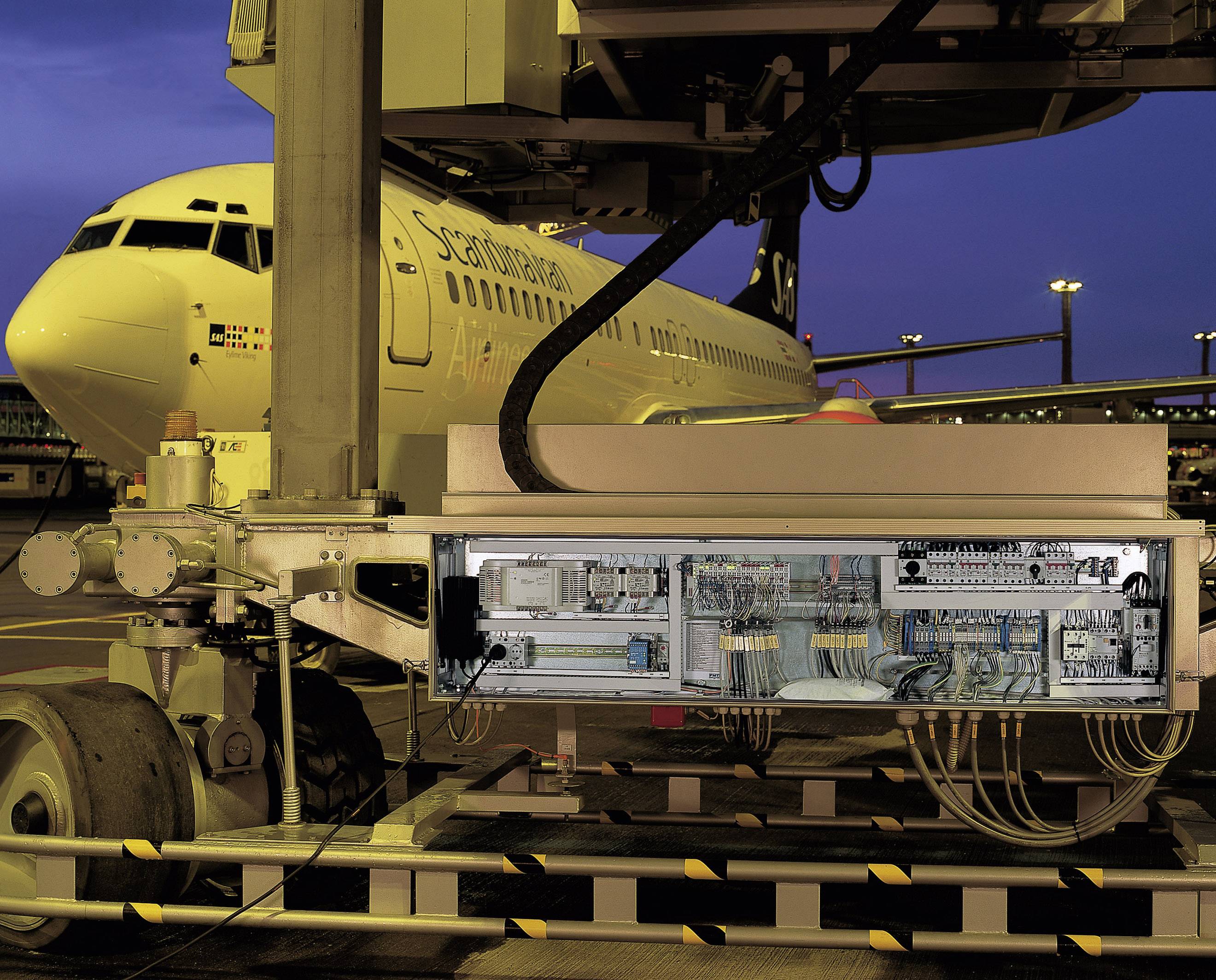 An aircraft is parked on the tarmac, while a service vehicle with its electronics cover open is connected for maintenance.