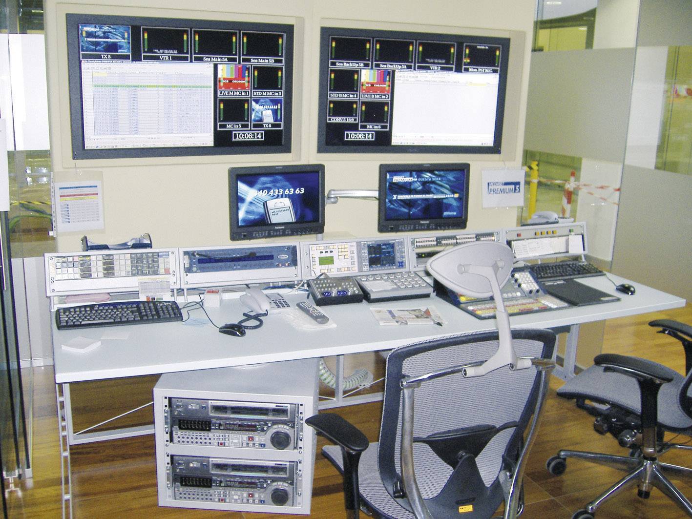 A control room with multiple screens, keyboards and technical devices placed on a desk. In the background, two large monitors are mounted on the wall.