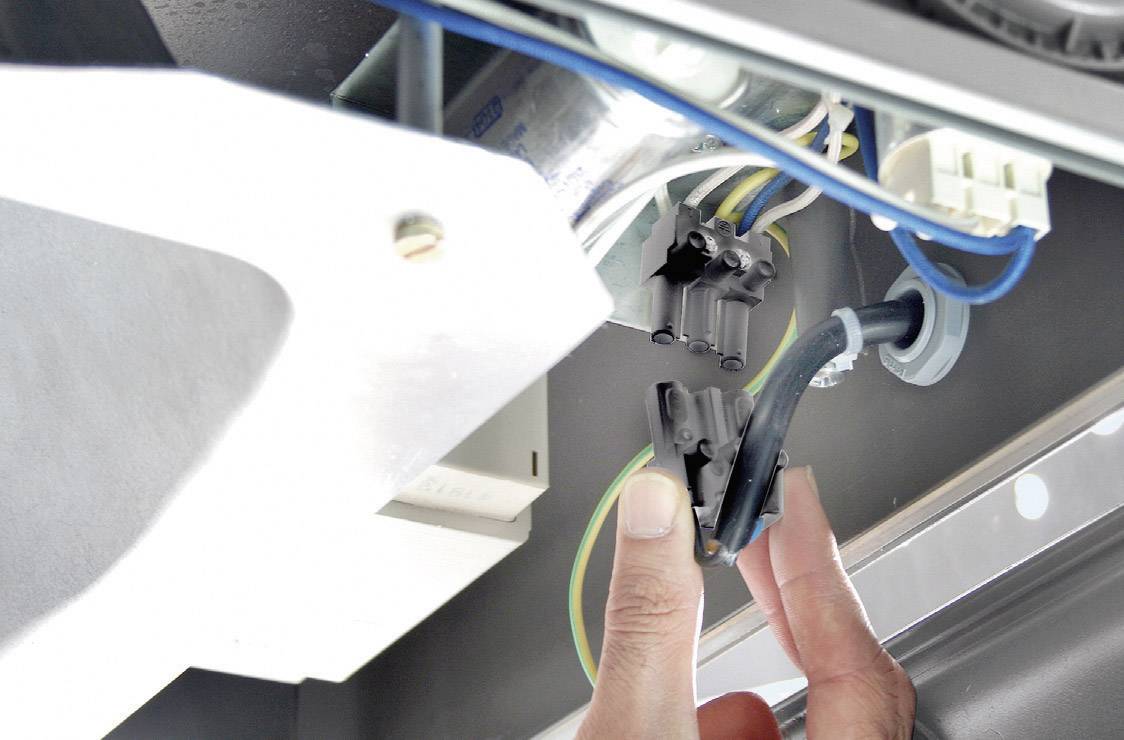 A person is pointing at a complex wiring arrangement beneath a device. Several cables and connections that are relevant to electronics are visible.