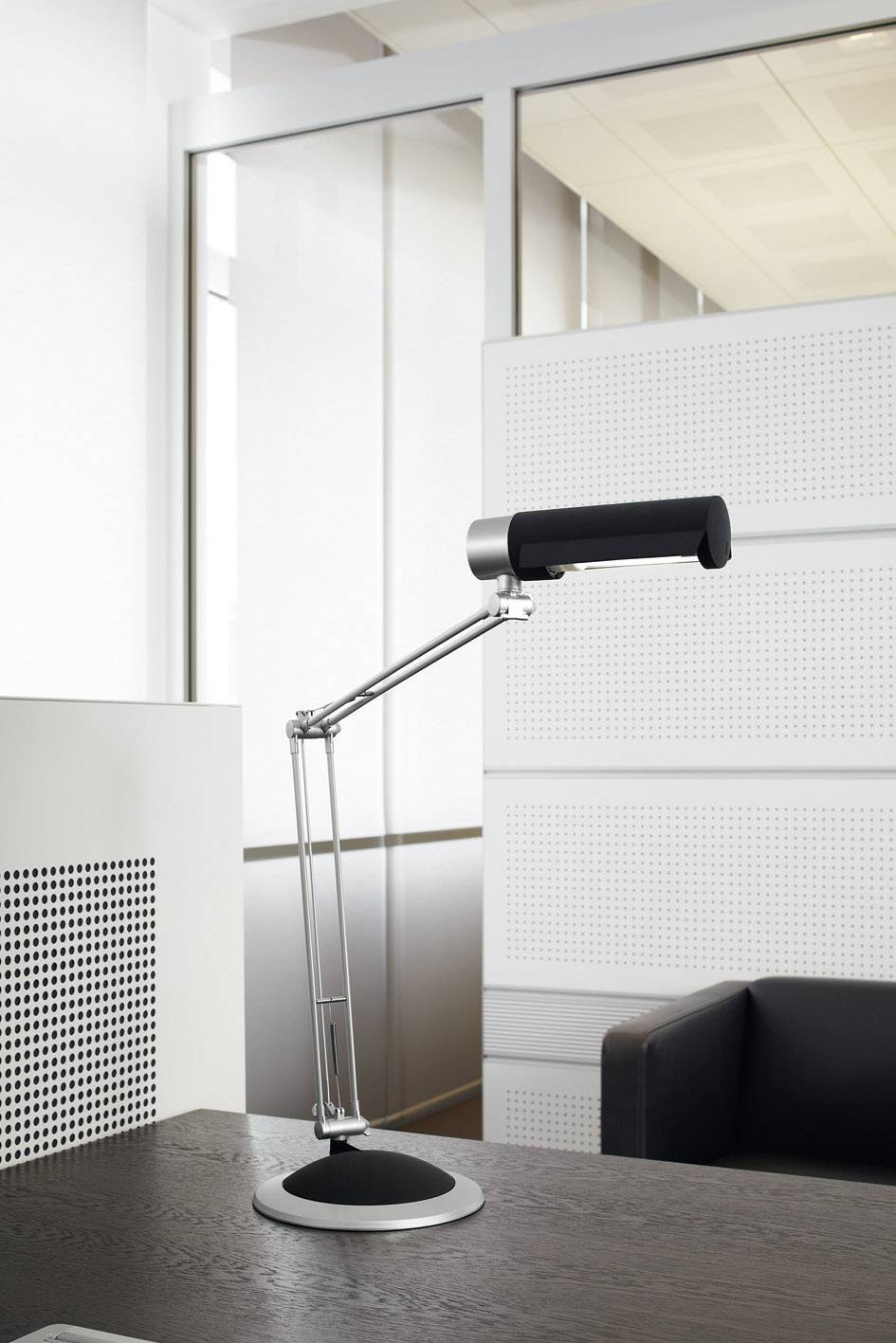 A modern desk lamp with a flexible arm sits on a dark wooden table in a brightly lit, contemporary office.