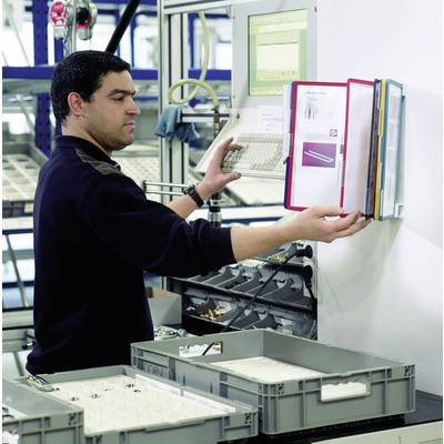 A man is working in a factory environment, operating a computer and checking documents in a file folder, in front of several storage boxes.
