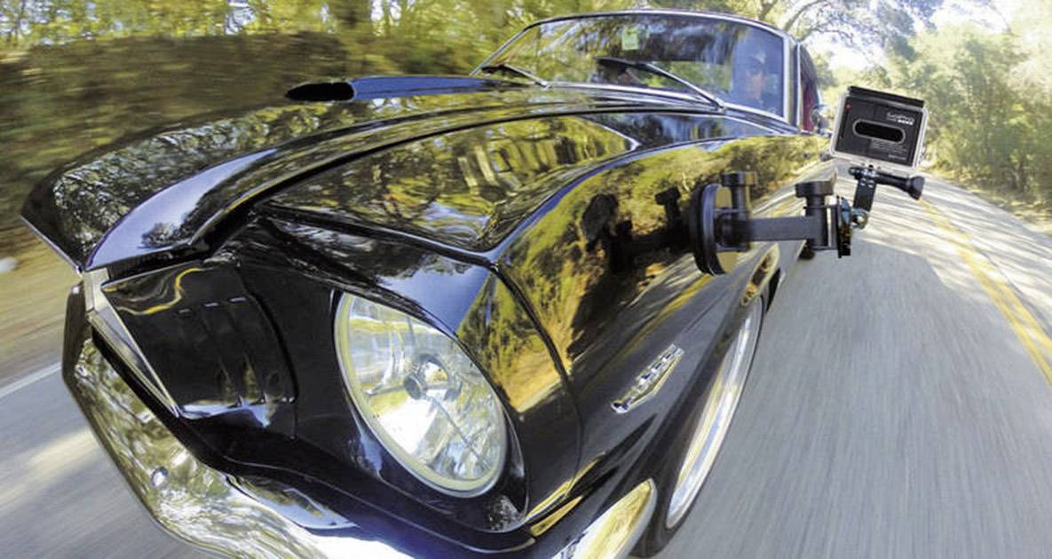 A black car is driving fast along a winding country road. A camera is mounted on the bonnet to record the journey.