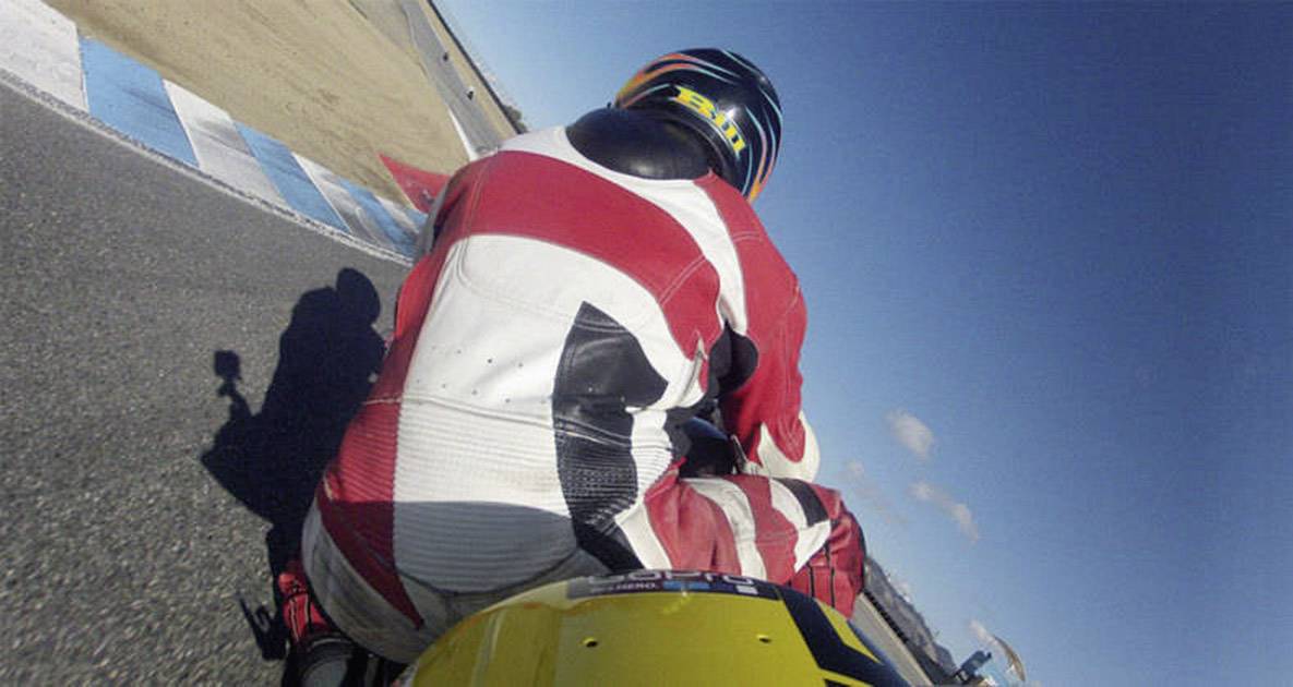 A motorcyclist in a red suit is riding through a bend on a racing circuit. A clear sky is visible in the background.