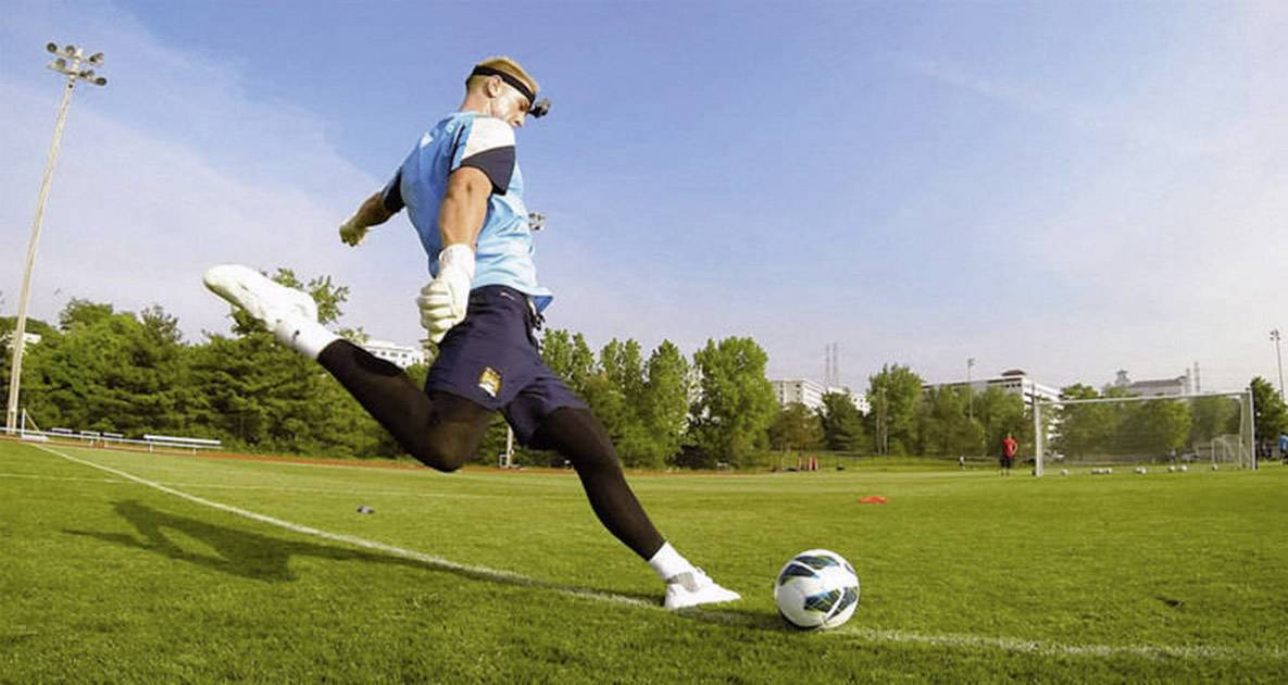 A football goalkeeper is practising on a green pitch. He is wearing a blue vest and black trousers and is kicking a ball with his right foot.