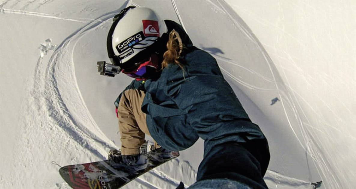 A person is snowboarding down a snow-covered slope. They are wearing a helmet with a camera.