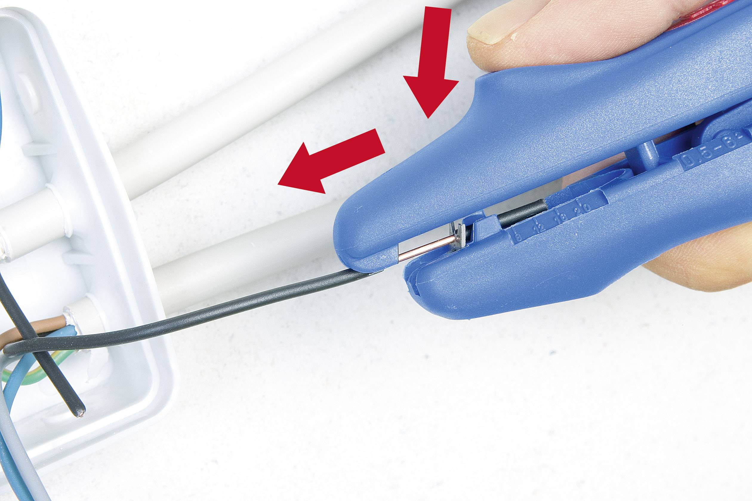 A hand is using a blue tool to strip the insulation from a black cable. Two red arrows point to the stripping point.