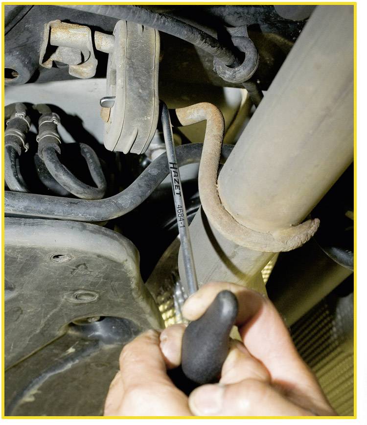 A person is using a tool to work on a component beneath a vehicle. The focus is on the hand holding a spanner.
