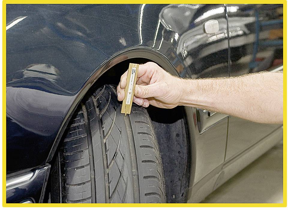 A hand measures the tread depth of a car tyre with a tread depth gauge. The car is navy blue.