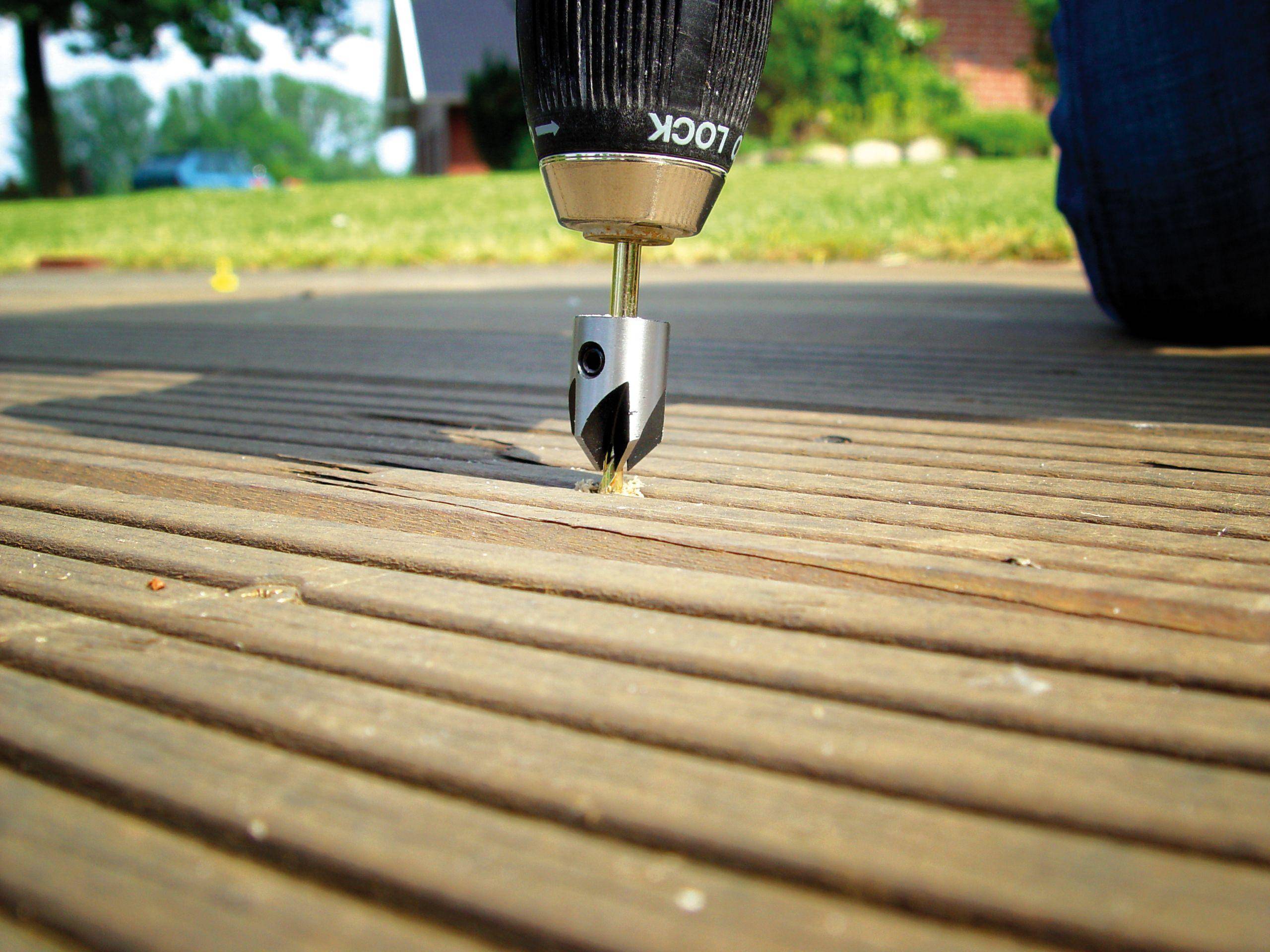 A drill is working on an outdoor wooden terrace. The drill bit is pressing small wood shavings out of the timber. Grass and trees are visible in the background.