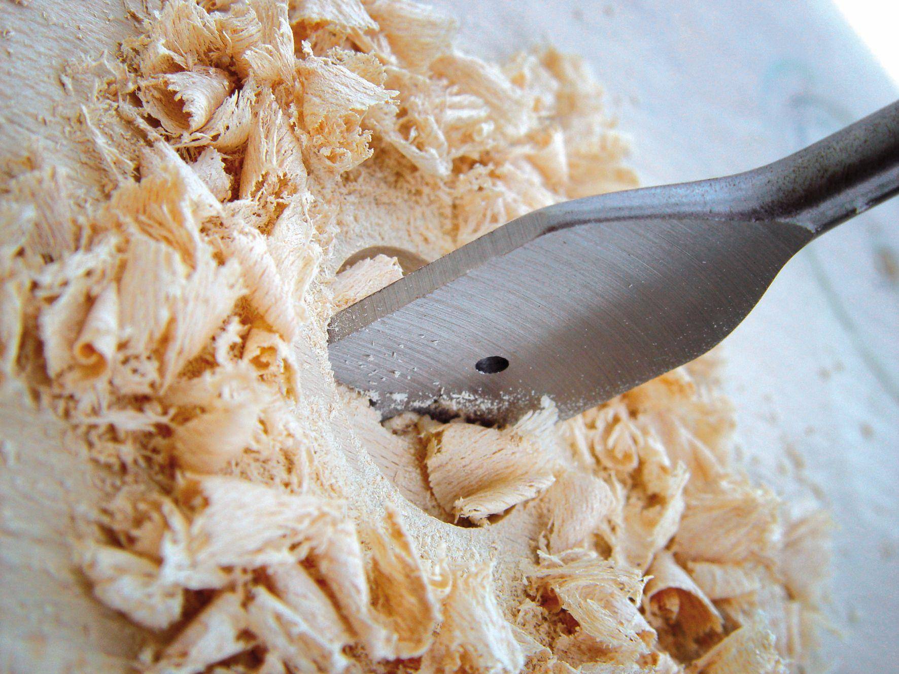 Close-up of a wood drill bit drilling a hole into a wooden board, surrounded by fresh wood shavings.