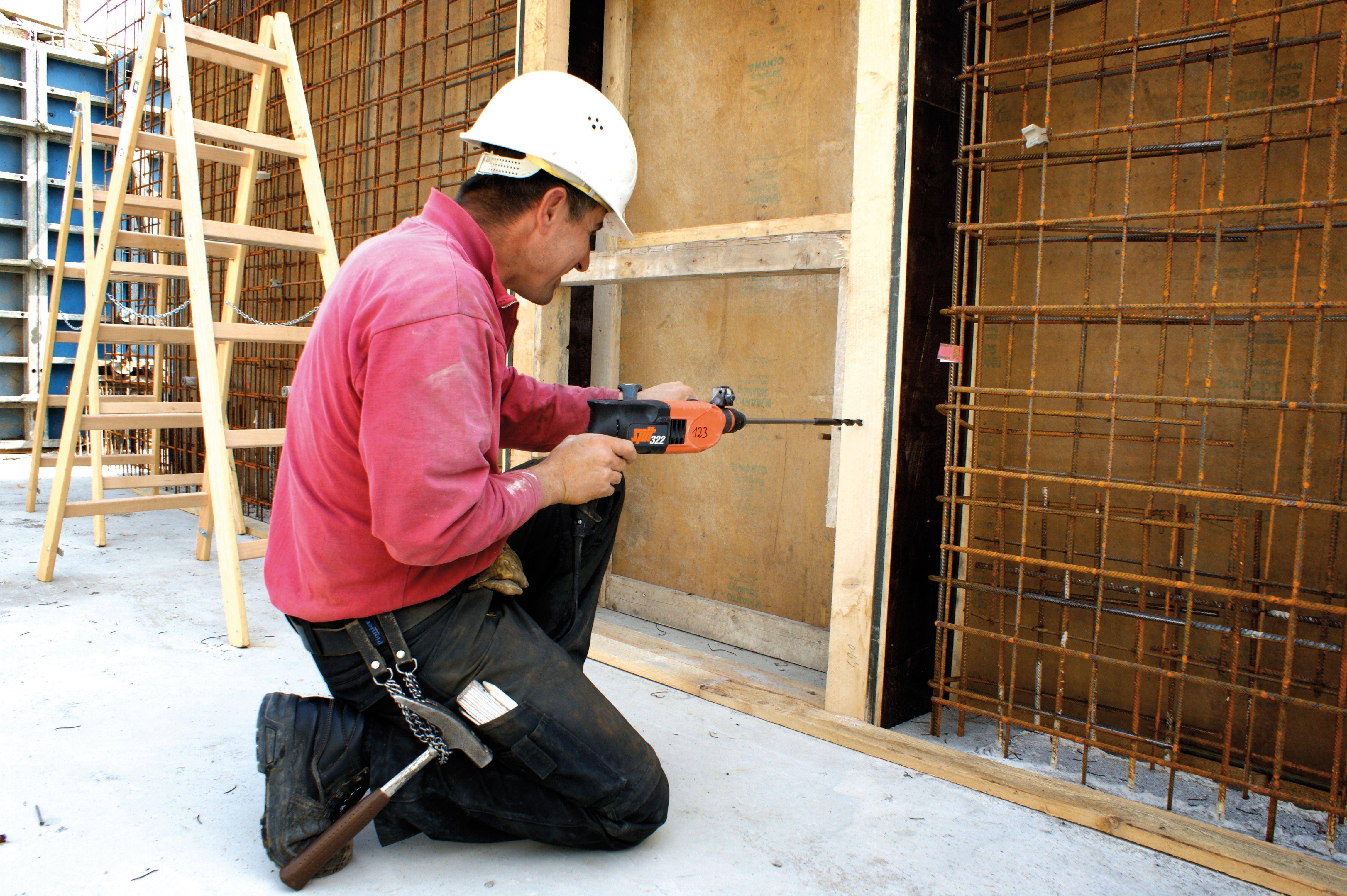 A construction worker is kneeling and drilling into a steel grid wall. He is wearing a white hard hat and a red shirt. A ladder is standing in the background.