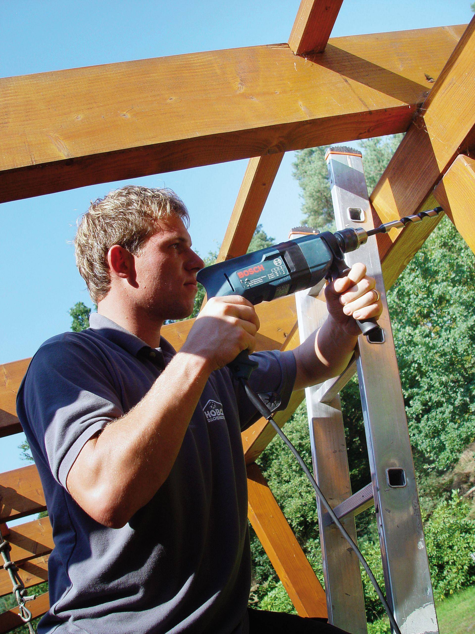 A man is standing on a ladder and using a drill to secure wooden boards to a frame outdoors.