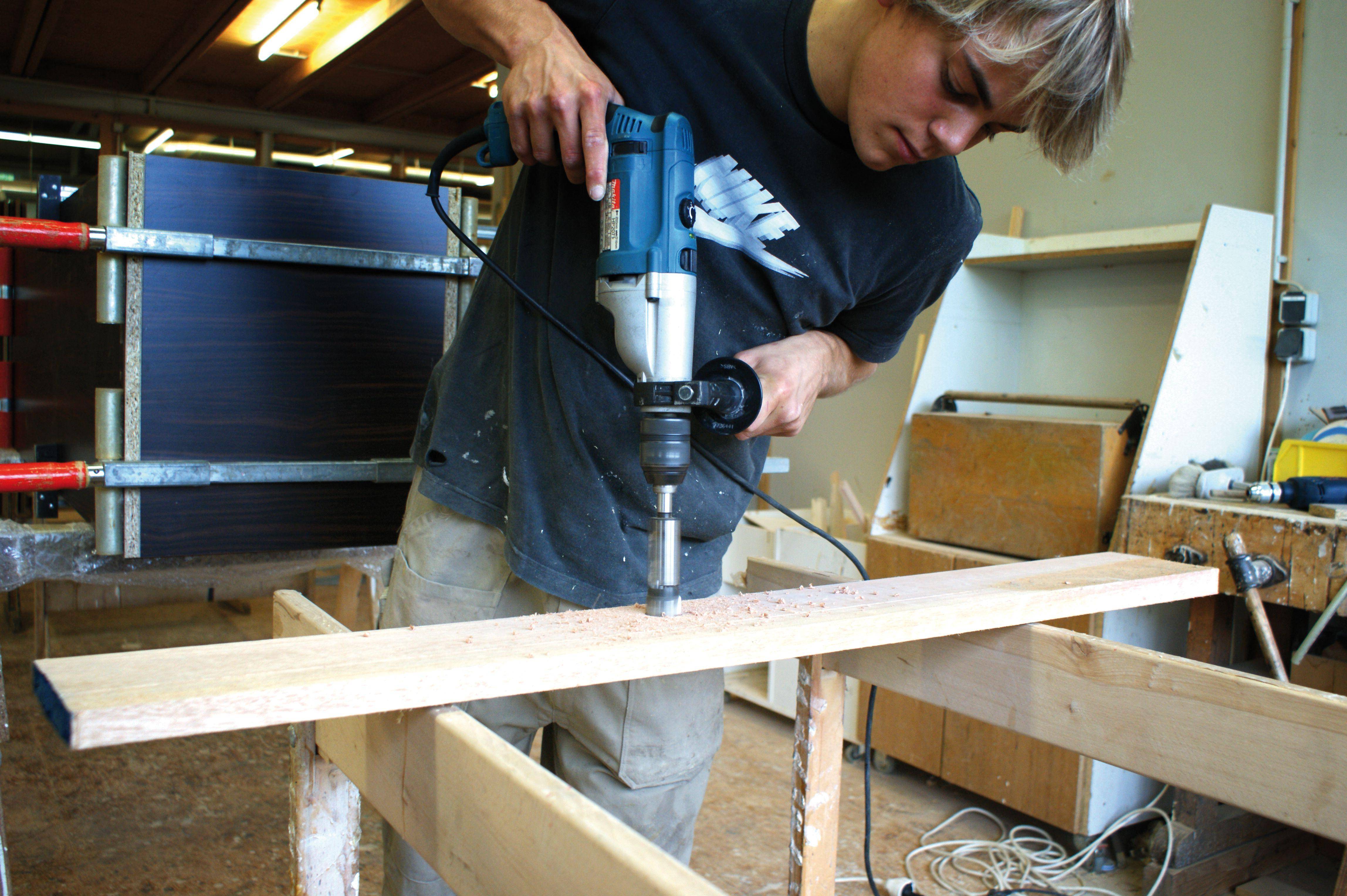 'A young man is drilling a hole in a wooden board with an electric drill in a workshop.'