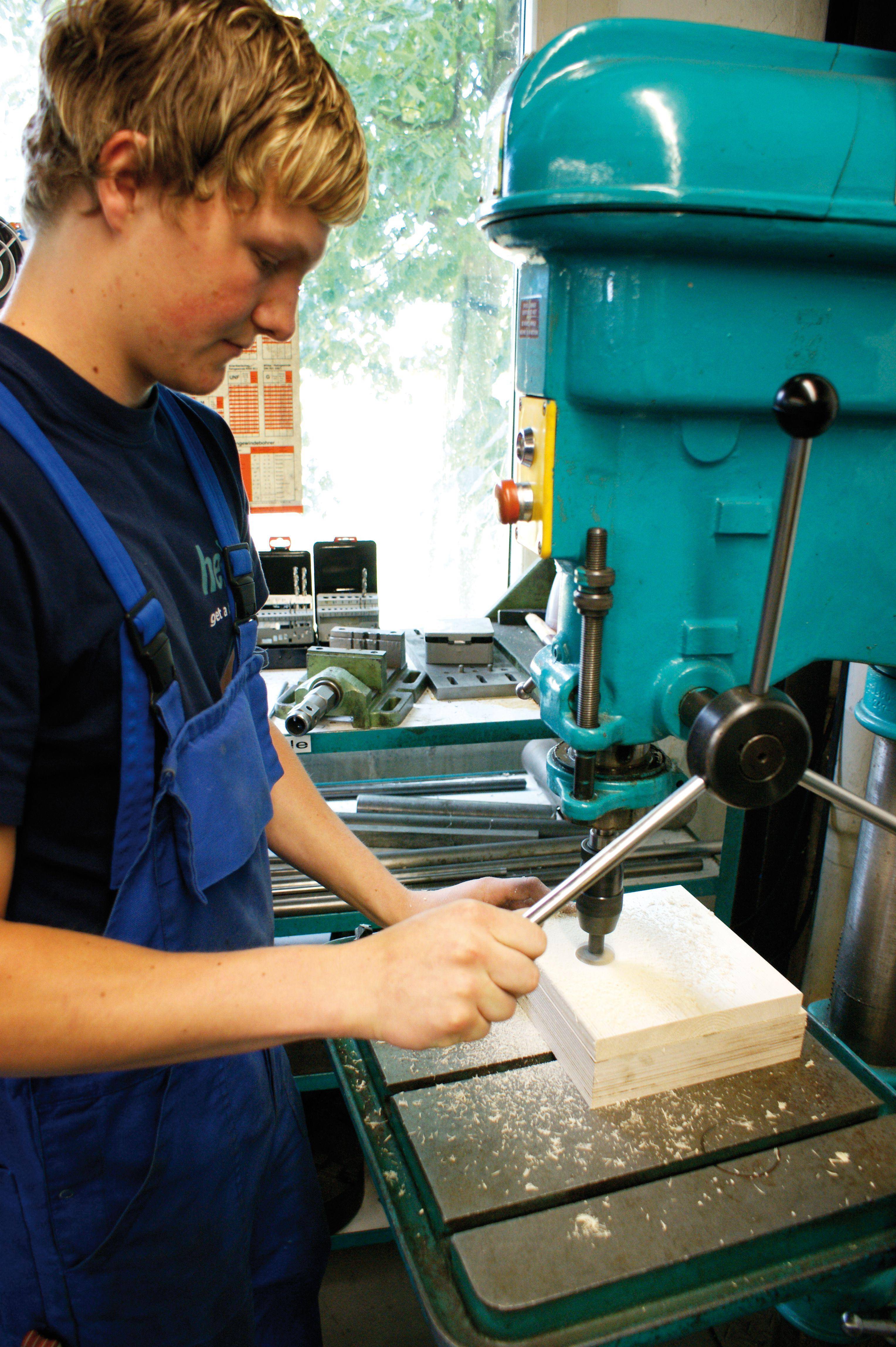 A young man in blue workwear is operating a drill to bore a hole into a piece of wood in a workshop.