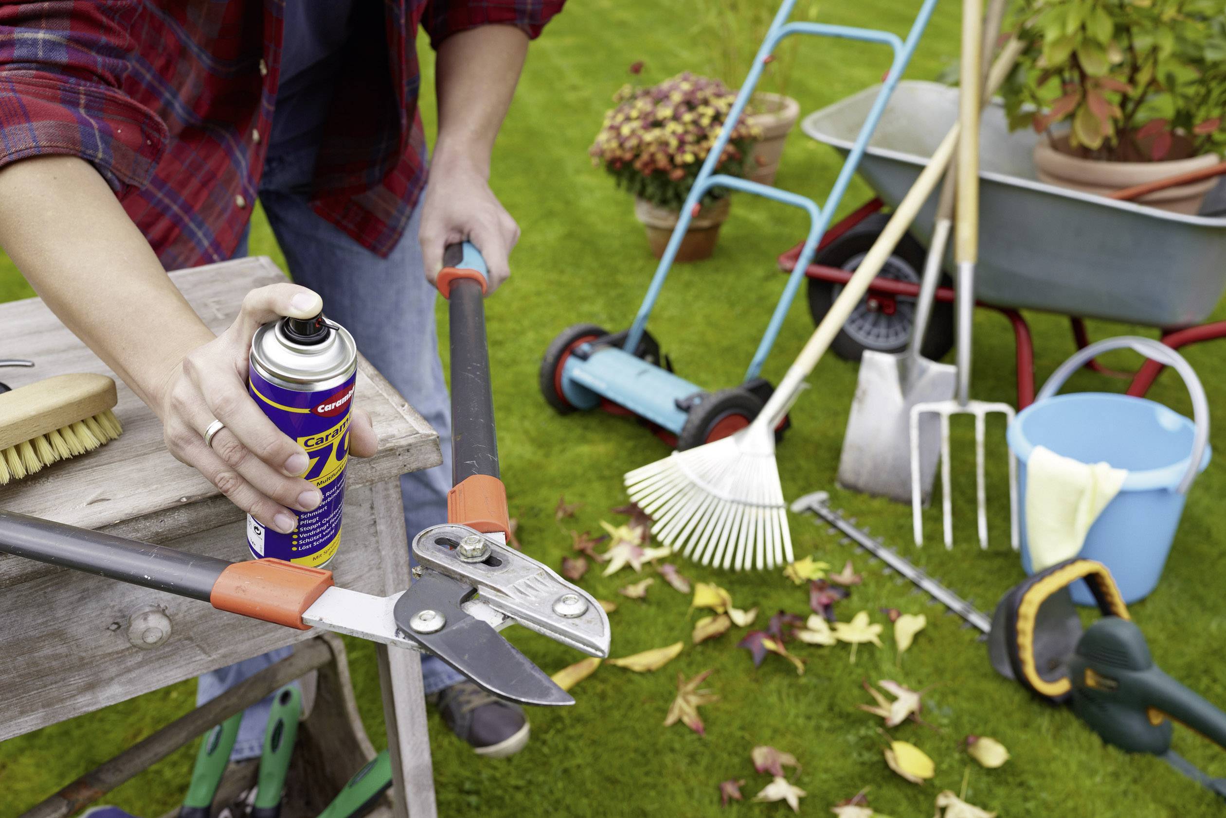 A person is lubricating gardening tools with a lubricant. Various garden implements and autumn leaves can be seen in the background.