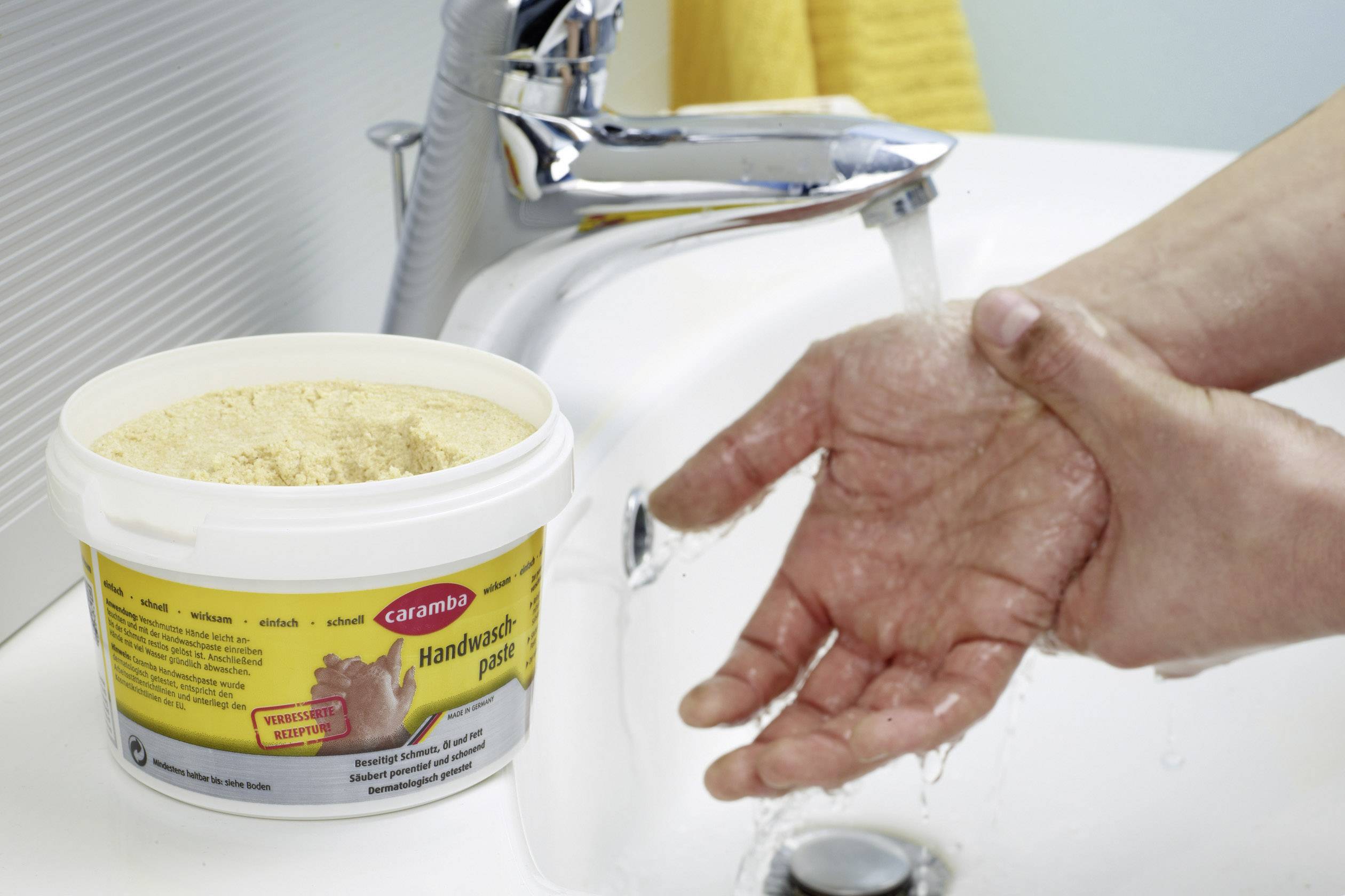 A person is washing their hands at a sink, with a bucket of 'hand wash paste' nearby. Running water flows from the tap, with a yellow hand towel in the background.