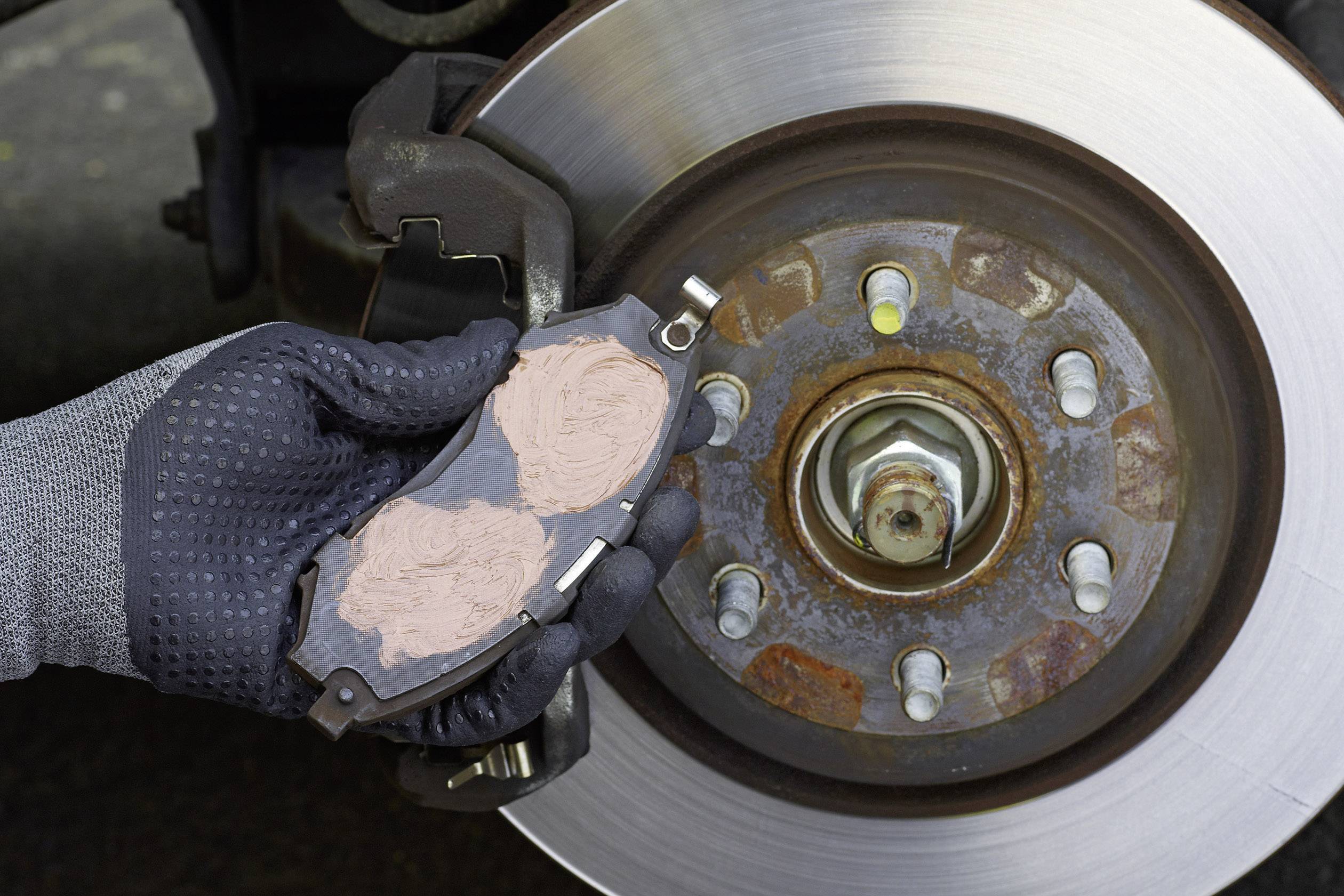 A hand in a glove holds a brake pad in front of a car braking system. The pad has a red coating.