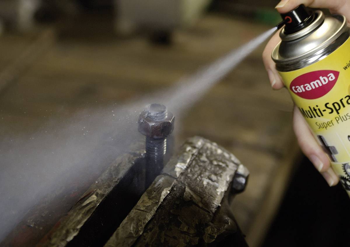 A person is spraying rust remover from a yellow can onto a rusted nut clamped in a vice in a workshop.