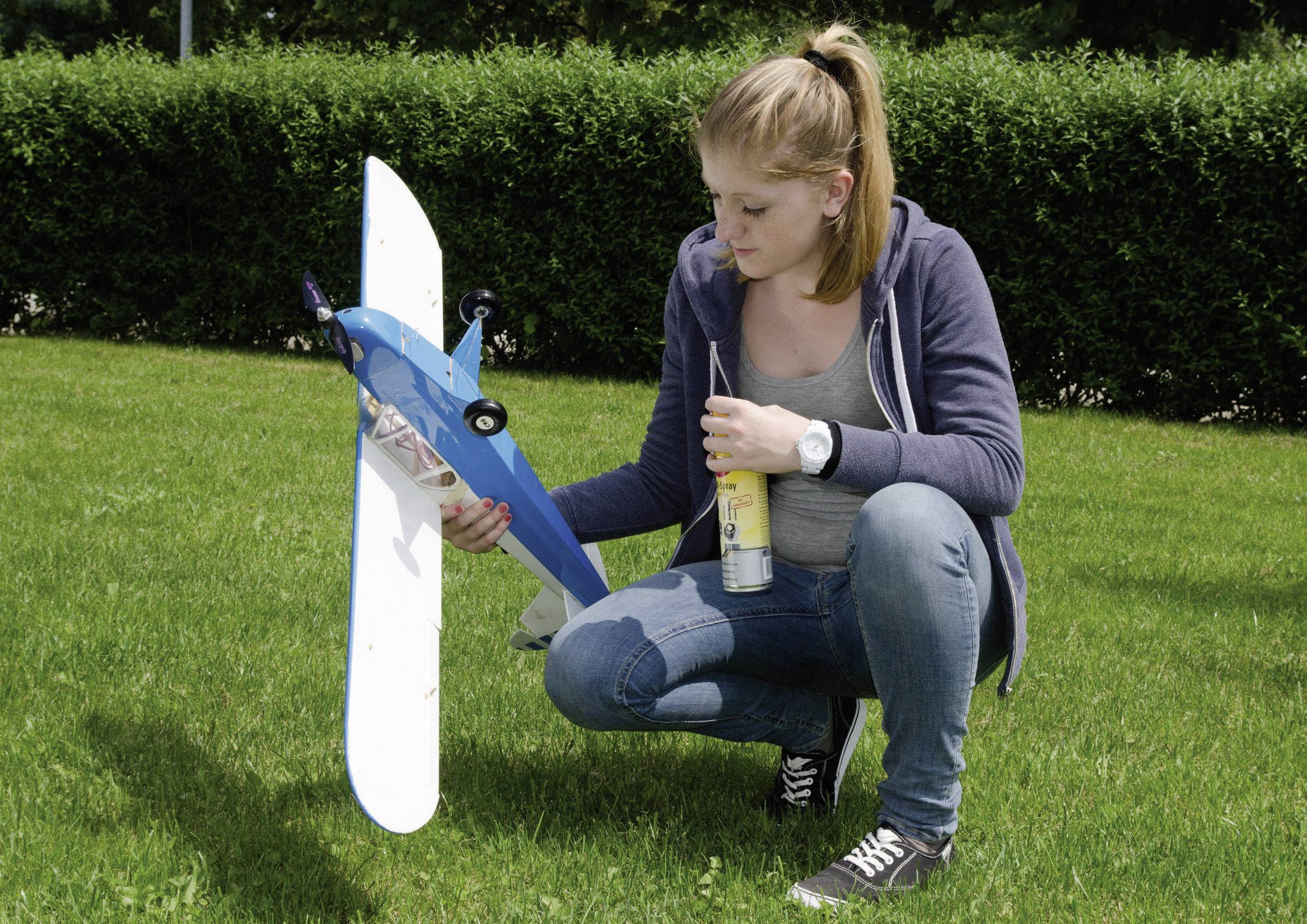 A woman kneels on a meadow, holding a model aeroplane in one hand and a spray can in the other.