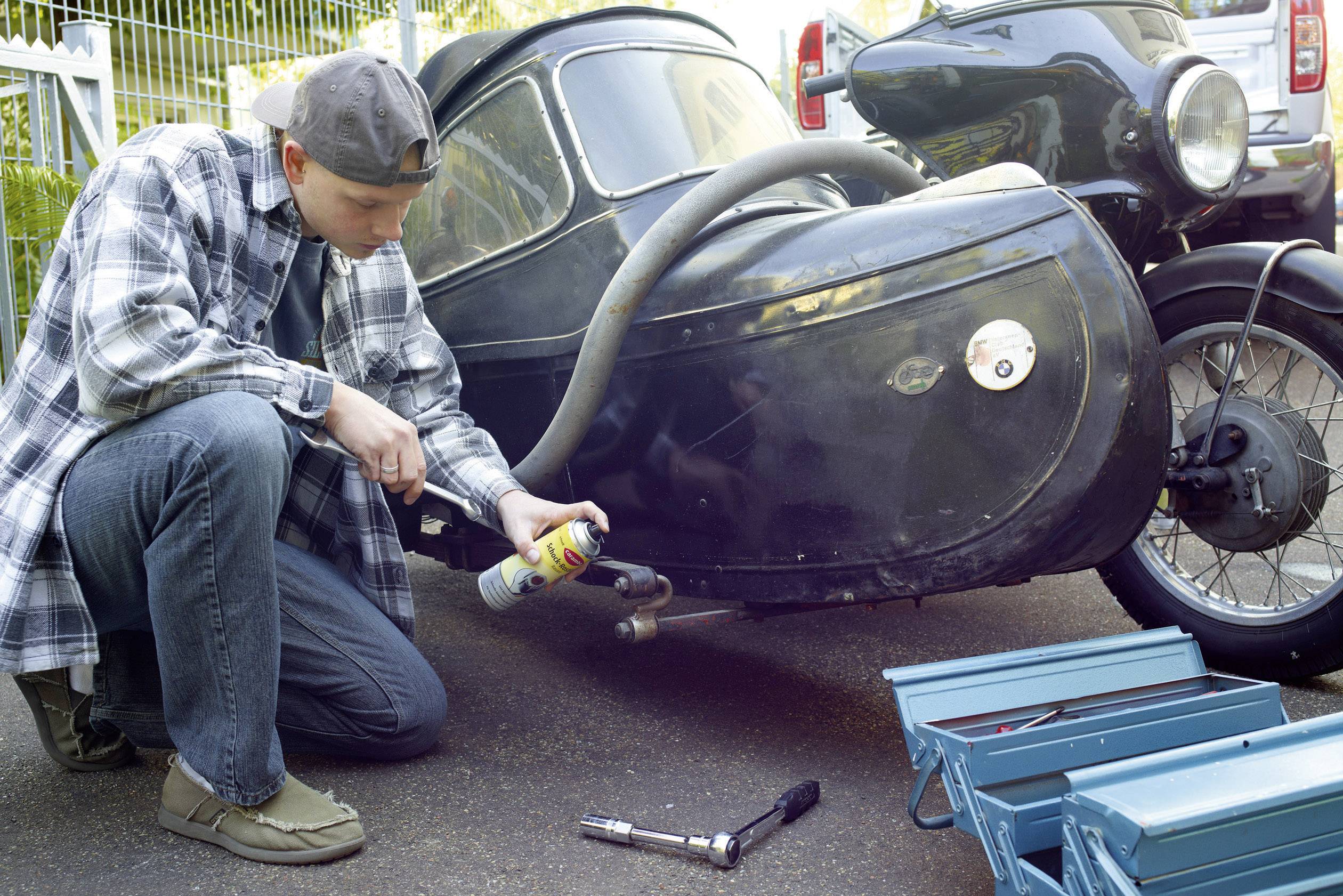 A man in a checked shirt and a backwards cap is kneeling beside a motorcycle with a sidecar and spraying lubricant onto parts of the sidecar.