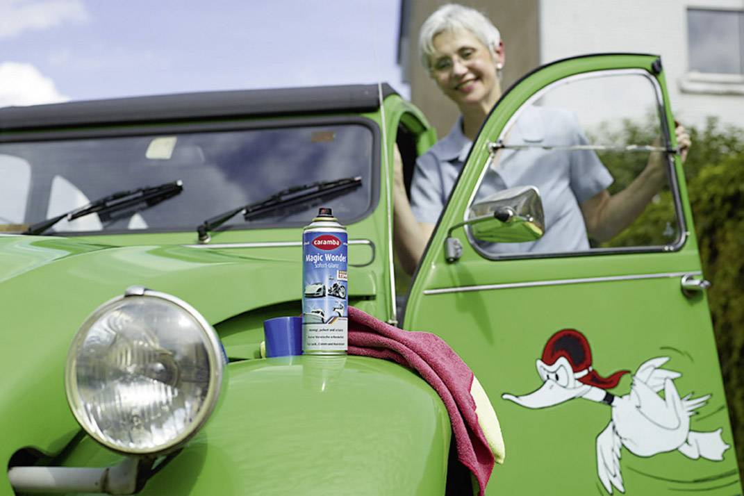 A person is standing smiling next to a green car with a duck sticker. A spray can of cleaning solution sits on the bonnet.