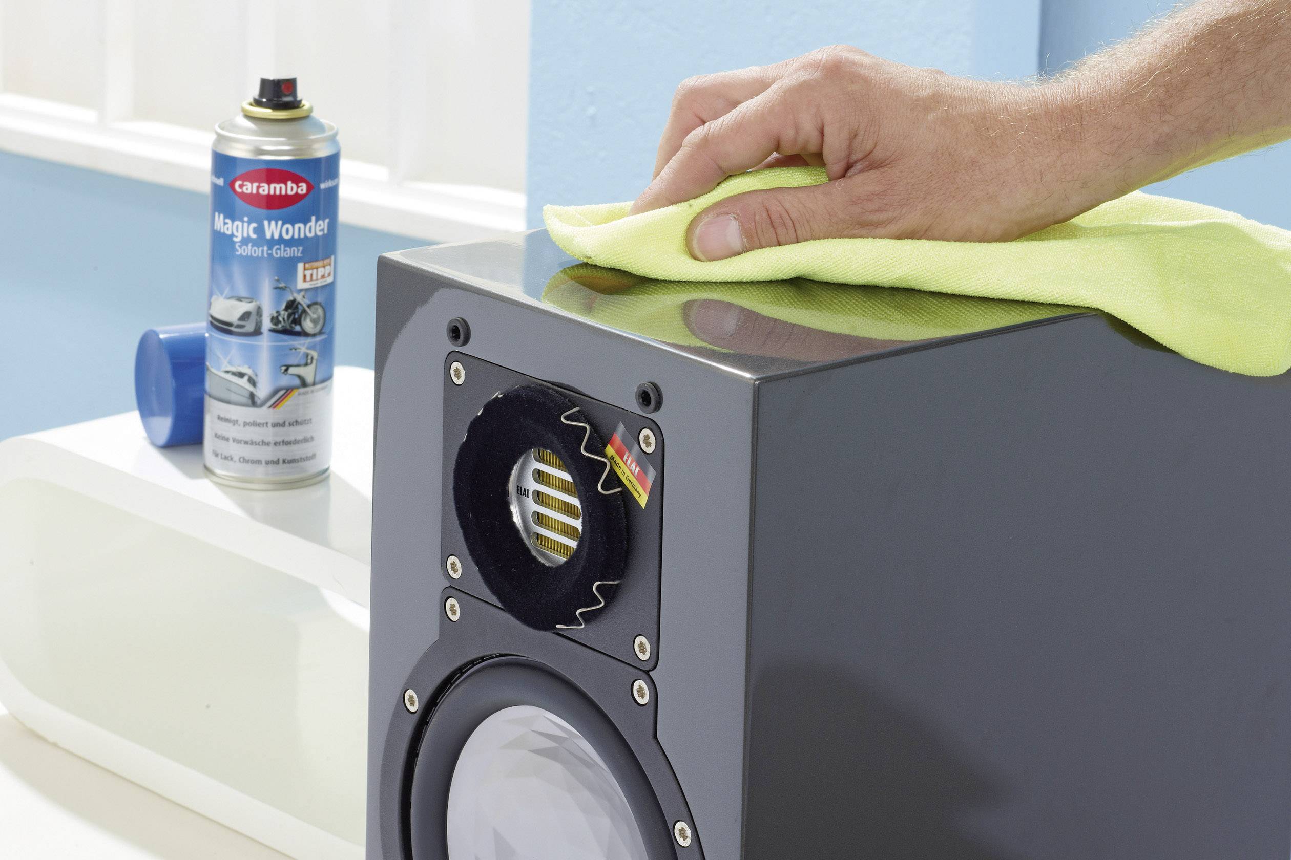 A hand wipes a speaker with a yellow cloth. In the background, a car care spray can sits on a white surface.