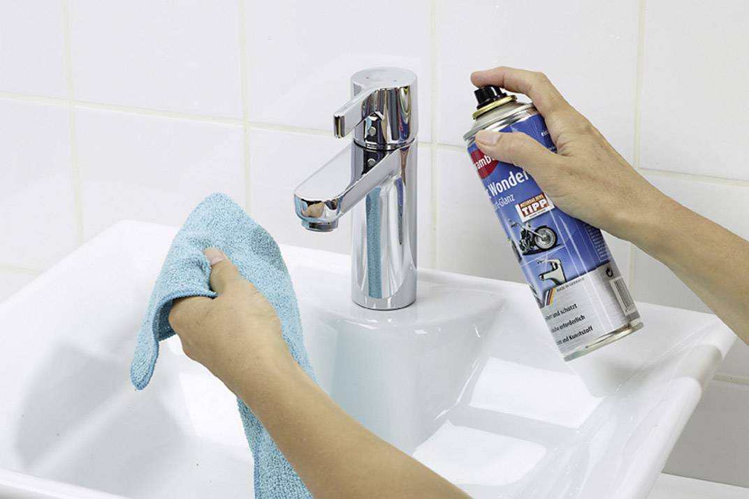 A person is cleaning a tap over a sink with a blue cleaning liquid and a cloth.
