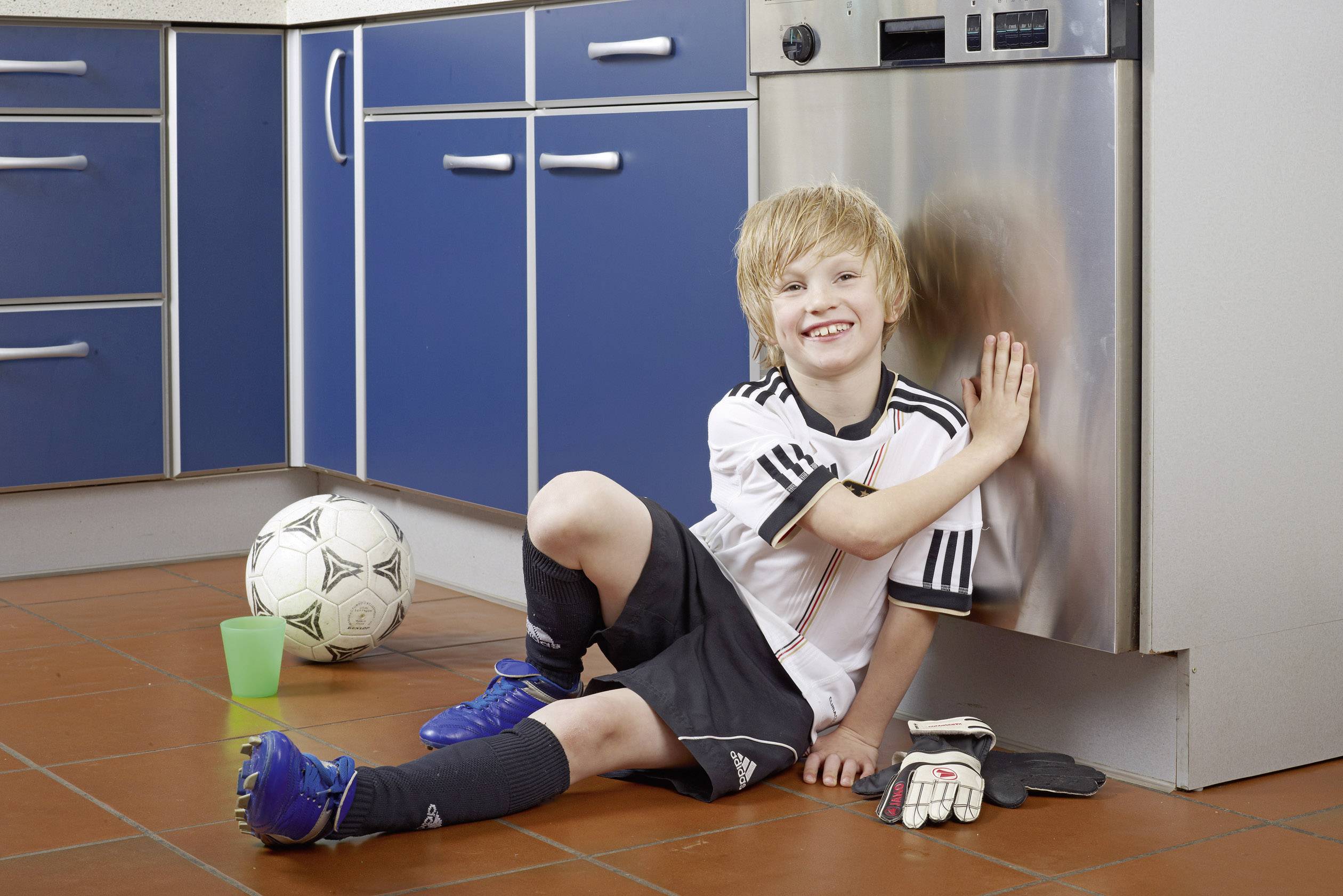 A boy in football kit sits on the kitchen floor, smiling and touching the dishwasher. Next to him is a football and a pair of goalkeeper's gloves.