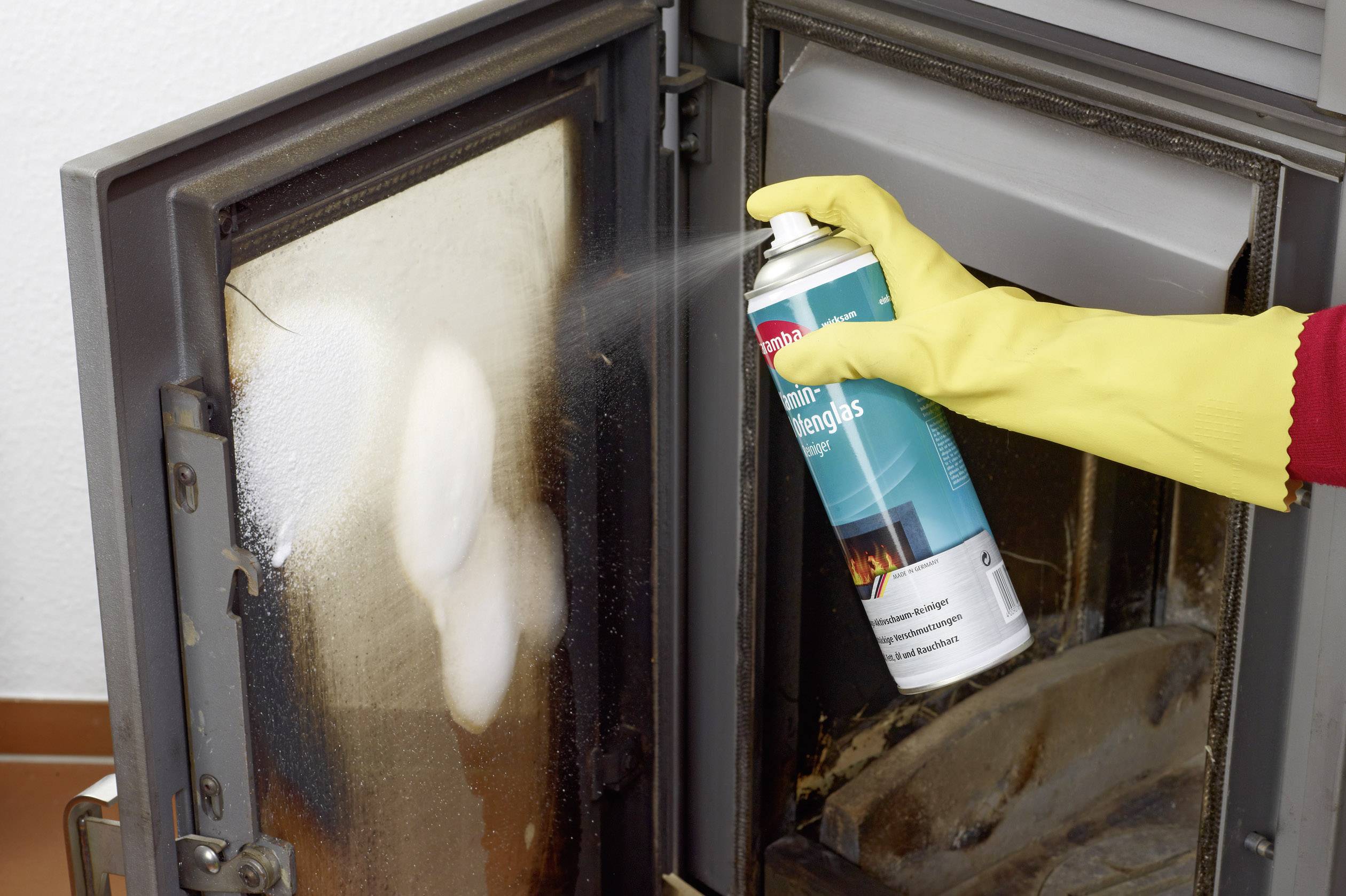 A hand wearing a yellow rubber glove sprays cleaner from a can onto the glass door of an oven.