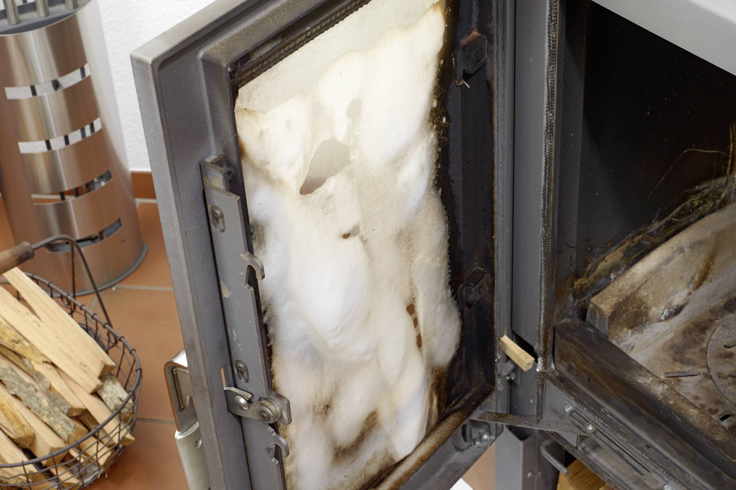 An open oven reveals the interior view with soot and ash. To the left of the oven stands a basket of chopped firewood.
