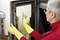 A person with short grey hair is cleaning the glass of an oven door with a blue cloth. They are wearing yellow rubber gloves.