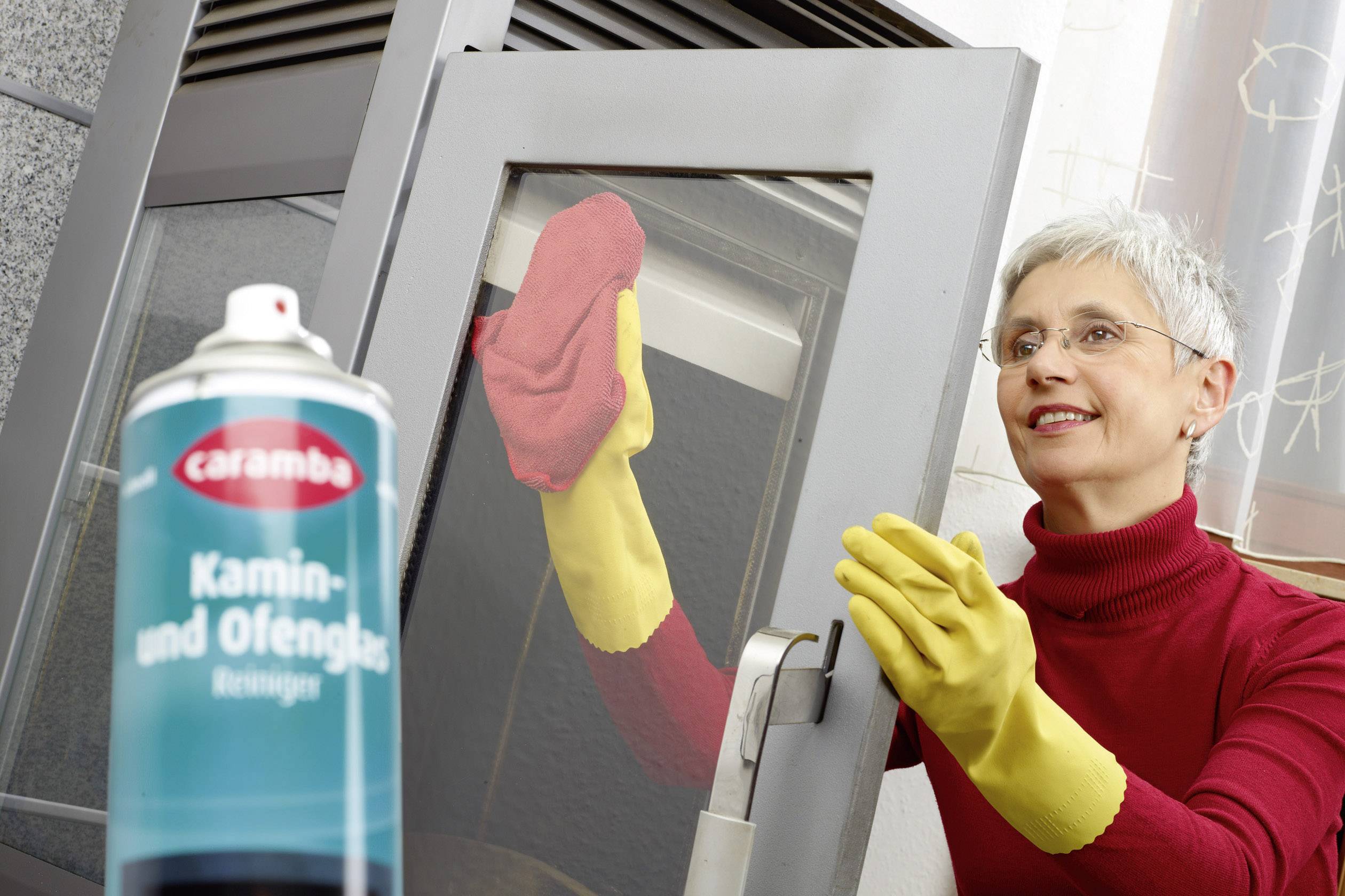 A woman wearing yellow gloves is cleaning a window with a red cloth. In the foreground is a tin of 'Fireplace and Oven Glass Cleaner'.