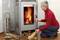 An elderly person lights a fireplace in the living room, next to a basket of chopped logs. They are wearing a red jumper and glasses.
