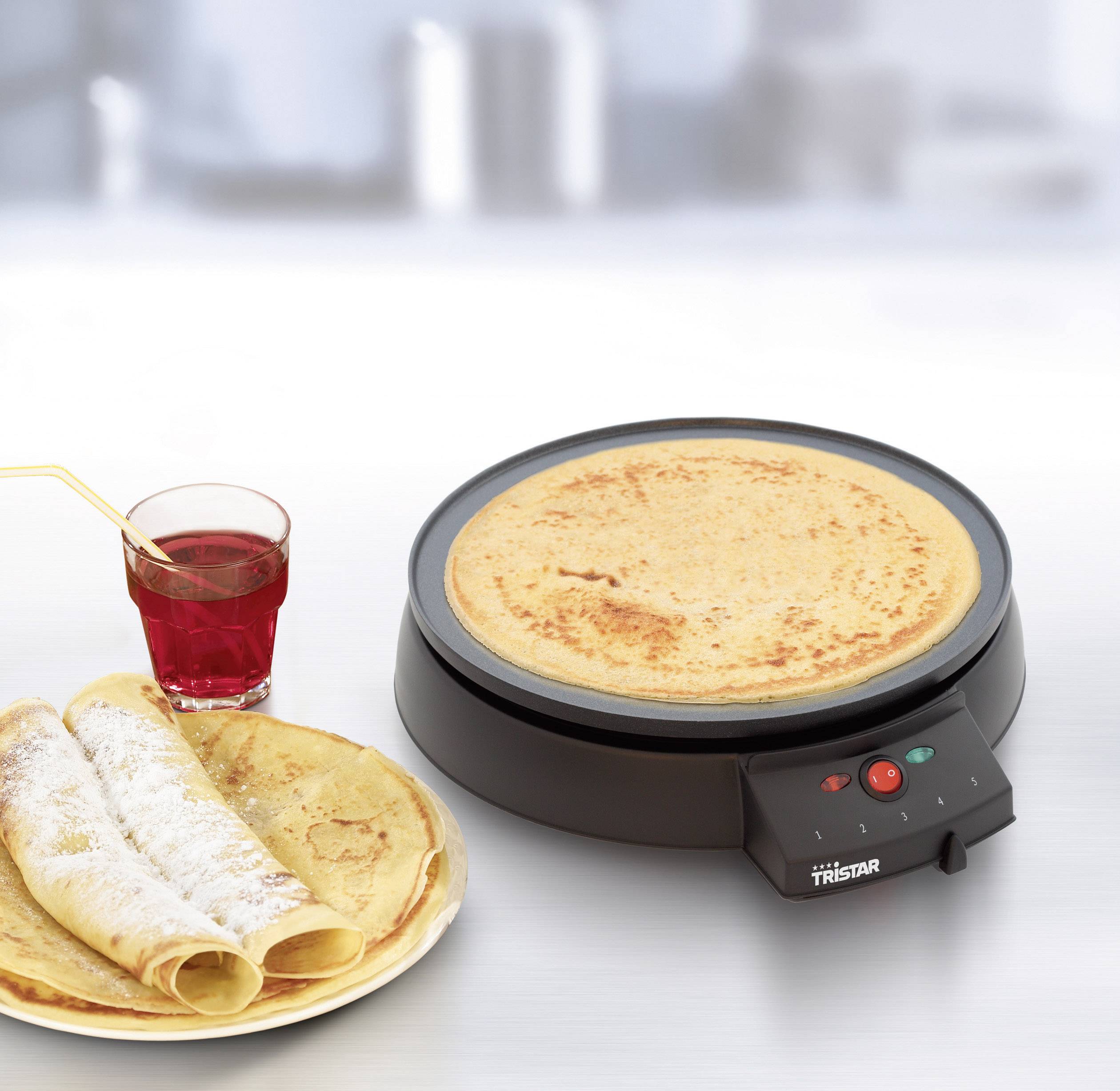 An electric crêpe maker with a freshly made crêpe beside it, along with a plate featuring two rolled crêpes and a glass of juice.