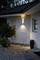 Two windows of a house with exterior lighting on the wall, illuminating a paved pathway and a small plant beside the façade.